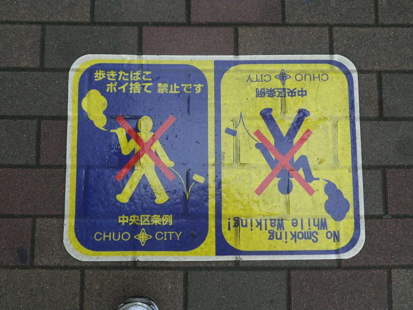 Yellow and blue Chuo City pavement sticker on brick paving showing a crossed-out walking smoker and the English text No Smoking while walking