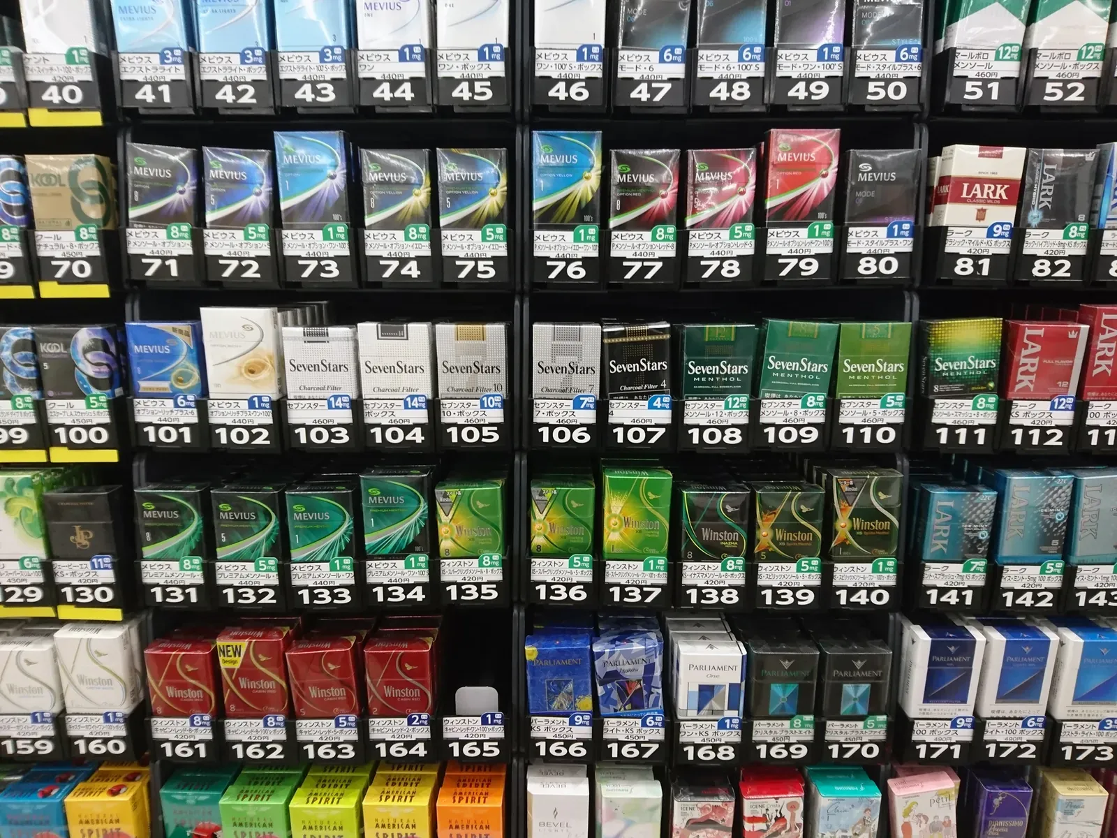 Nahaufnahme einer Zigarettenauslage in einem Convenience-Store mit Packungen von Mevius, Seven Stars, Winston und Lark, jeweils mit eigener Nummer und Preisschild