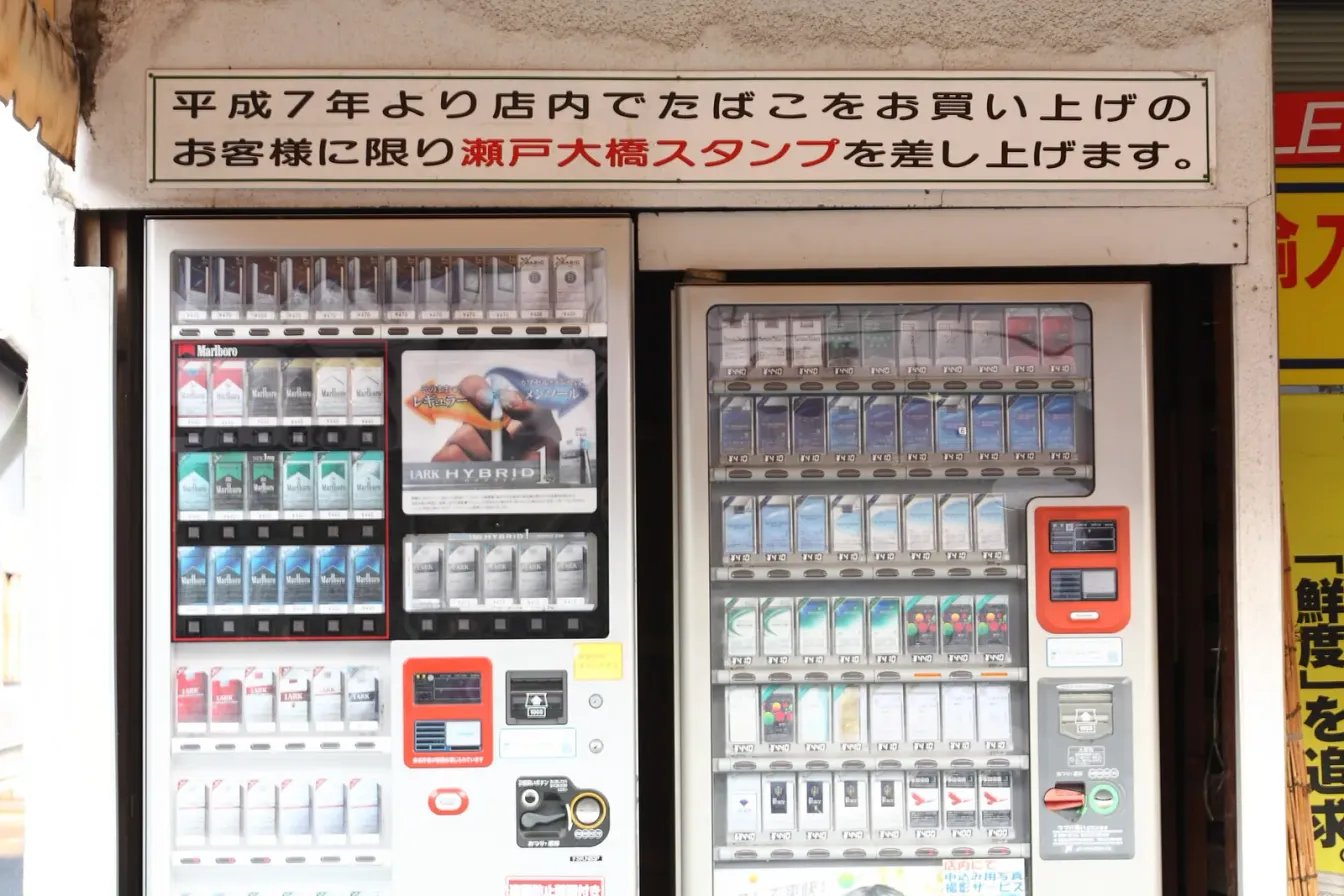 两台嵌在店面中的日本香烟自动售货机，摆有 Marlboro、Lark、Seven Stars 和 Hope 香烟，每台均配有红色 taspo 年龄验证读卡器