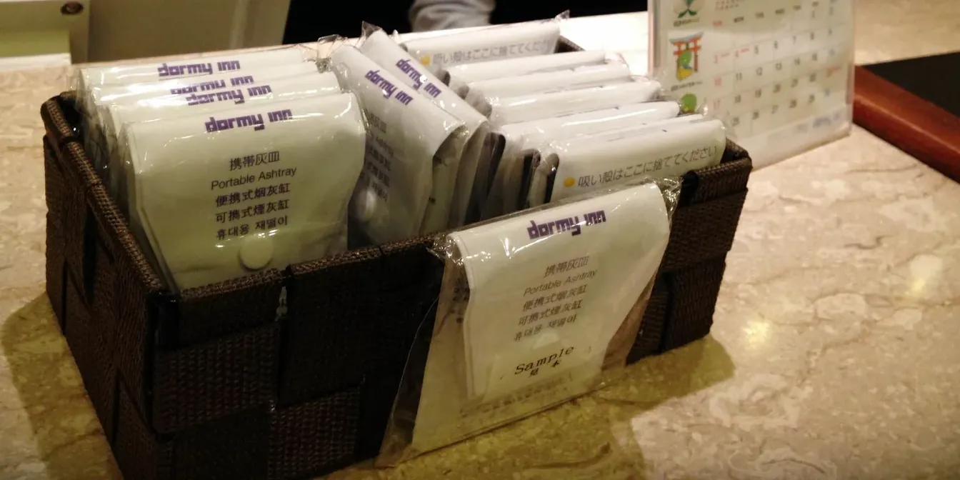 Ein Korb mit verpackten japanischen Taschenaschenbechern auf der Rezeption eines Hotels in Japan, beschriftet mit 携帯灰皿 / Portable Ashtray
