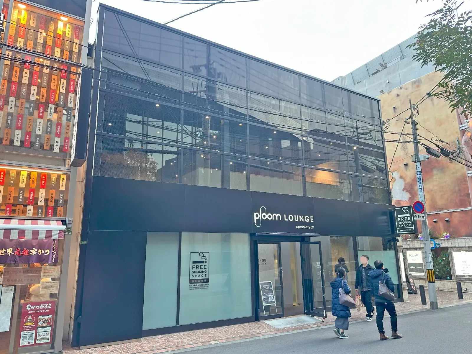 Fassade der Ploom Lounge in Osaka, Japan, mit einem Schild „Free Smoking Space“ im Fenster und vorbeigehenden Fußgängern
