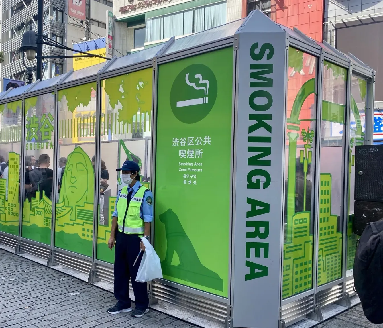 Öffentlicher Raucherbereich des Bezirks Shibuya mit Glaswänden an einer Straße in Tokio, mit den Aufschriften SMOKING AREA und 渋谷区公共喫煙所 in fünf Sprachen; innen rauchen Menschen, außen steht eine Aufsichtsperson