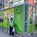东京街头一处由玻璃围板围起的涩谷区公共吸烟处，标有五种语言的“SMOKING AREA”和“渋谷区公共喫煙所”；有人在内吸烟，一名工作人员站在外面