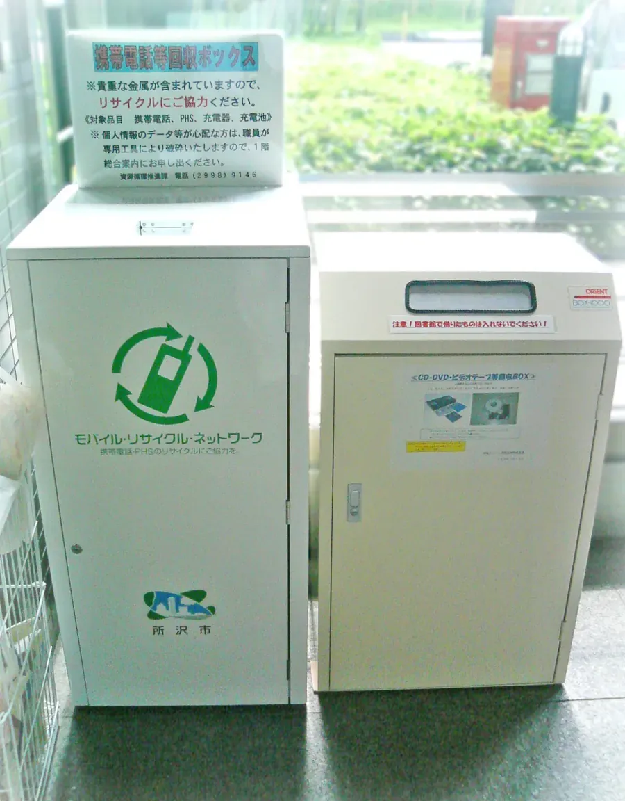 Small-electronics recycling collection box inside the entrance of a Japanese city office, labelled 携帯電話等回収ボックス for phones, chargers and rechargeable batteries