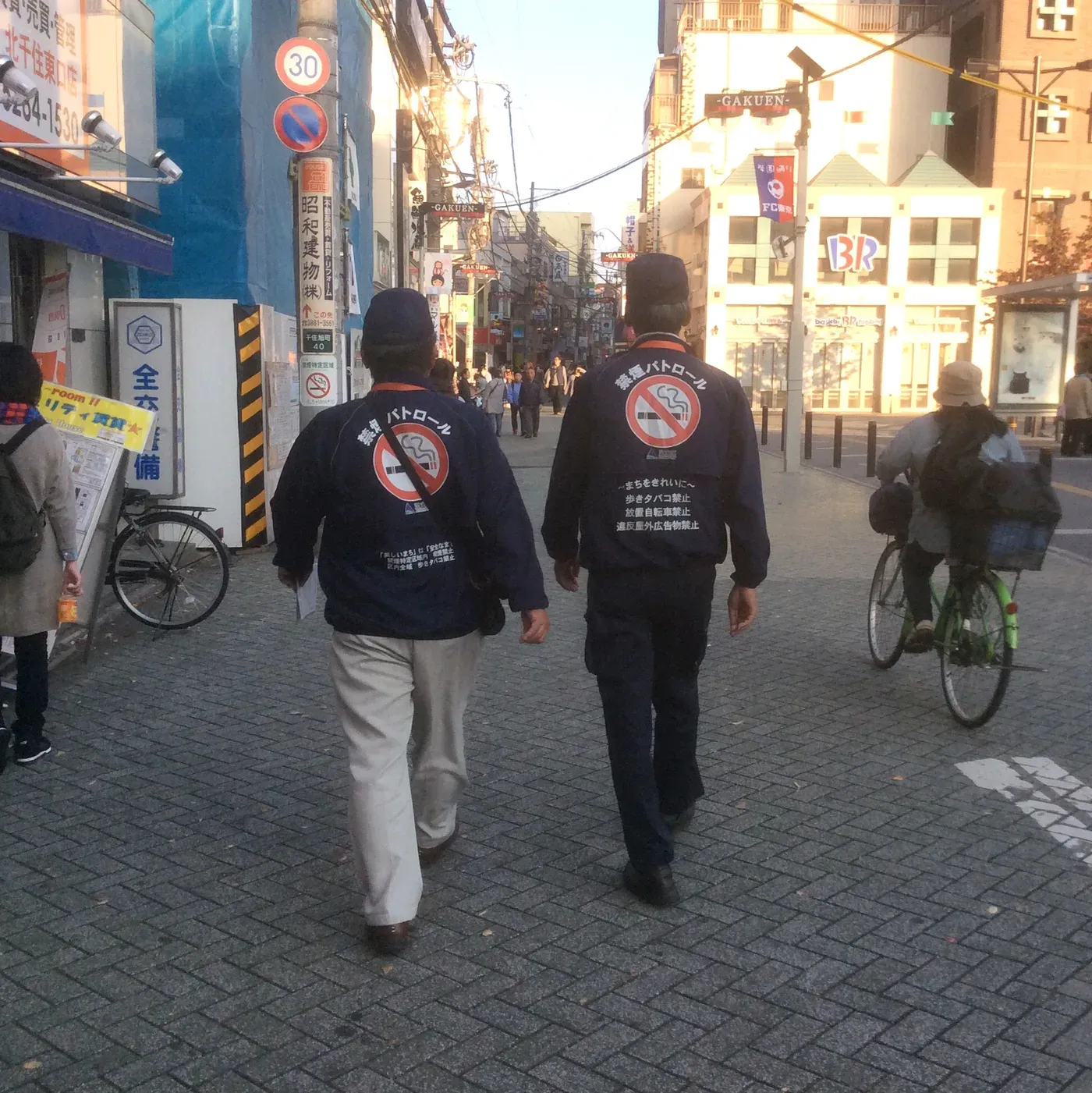 Two street smoking patrol officers in navy jackets printed with a crossed-out cigarette walking along a shopping street in Kita-Senju, Tokyo