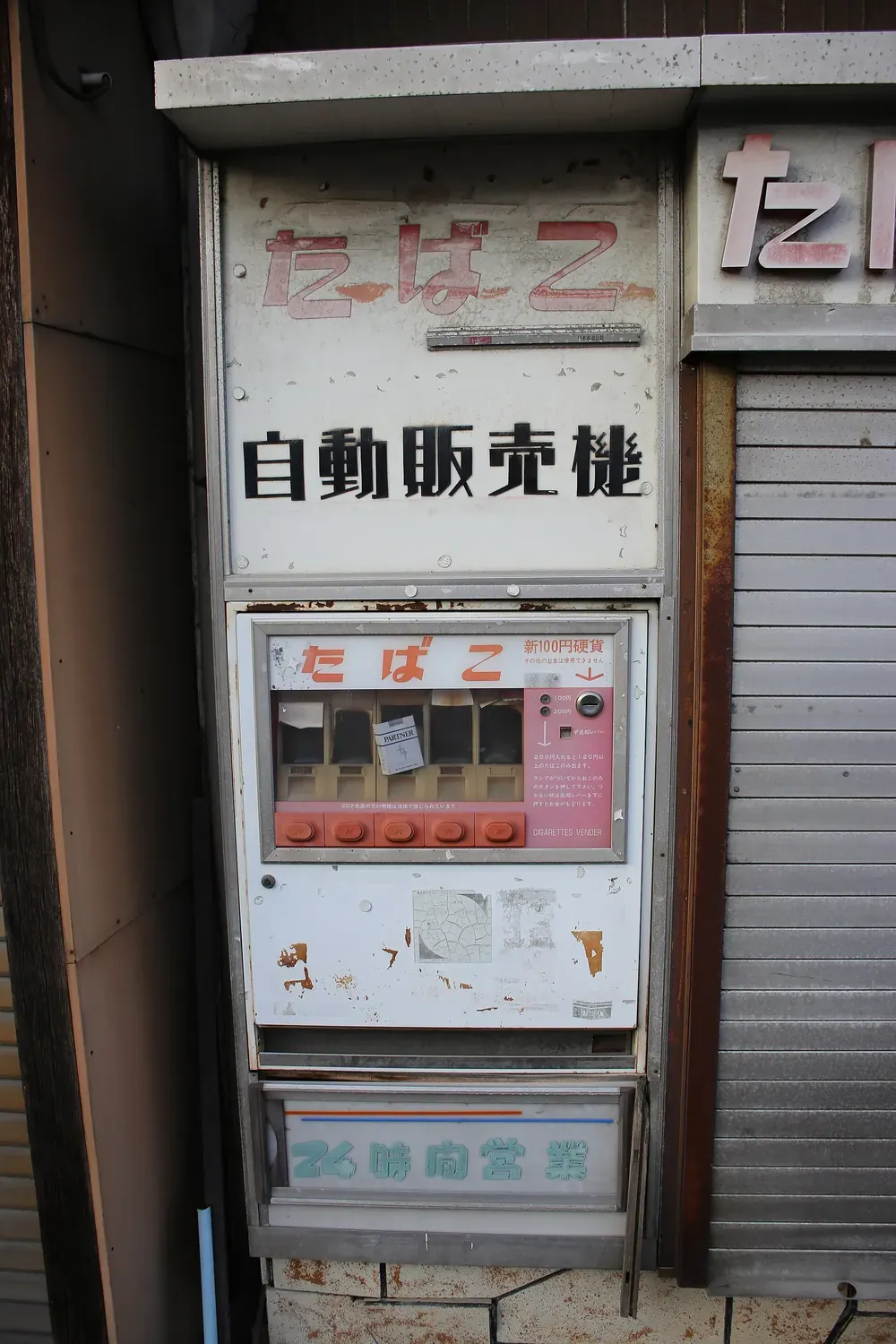 Ein verwitterter, rostender Zigarettenautomat vor einem geschlossenen japanischen Geschäft mit verblichener Beschilderung und abblätternder Farbe