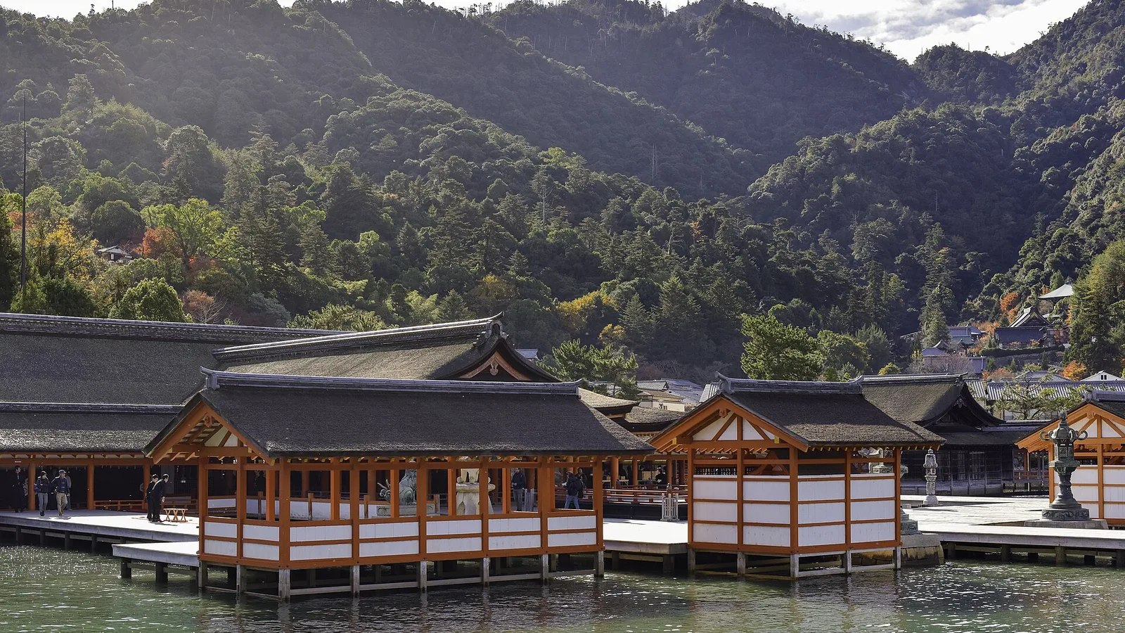 The vermilion halls of Itsukushima Shrine standing on stilts over the water at Miyajima, with forested hills behind