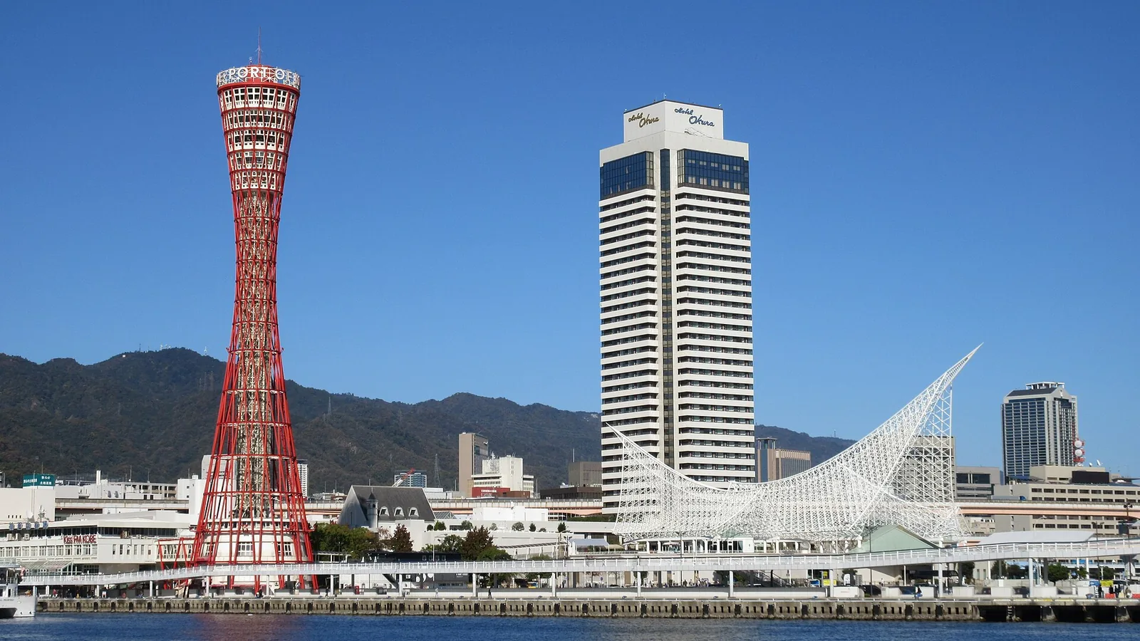 The red lattice of Kobe Port Tower beside the white sail roof of the Maritime Museum, seen across the harbour under a clear sky