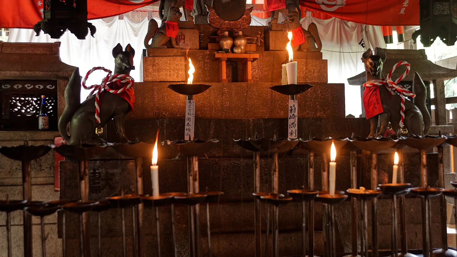 Candles burning before a stone altar at Fushimi Inari-taisha, flanked by bronze fox statues in red votive bibs