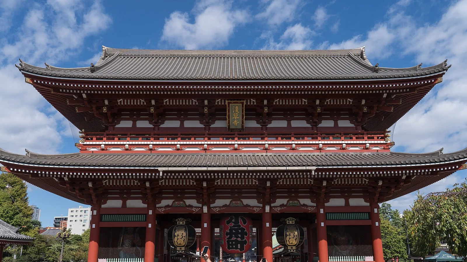 Sensō-ji's Hōzōmon gate in Asakusa under a blue sky, its two tiled roofs above three red pillars and a pair of giant paper lanterns