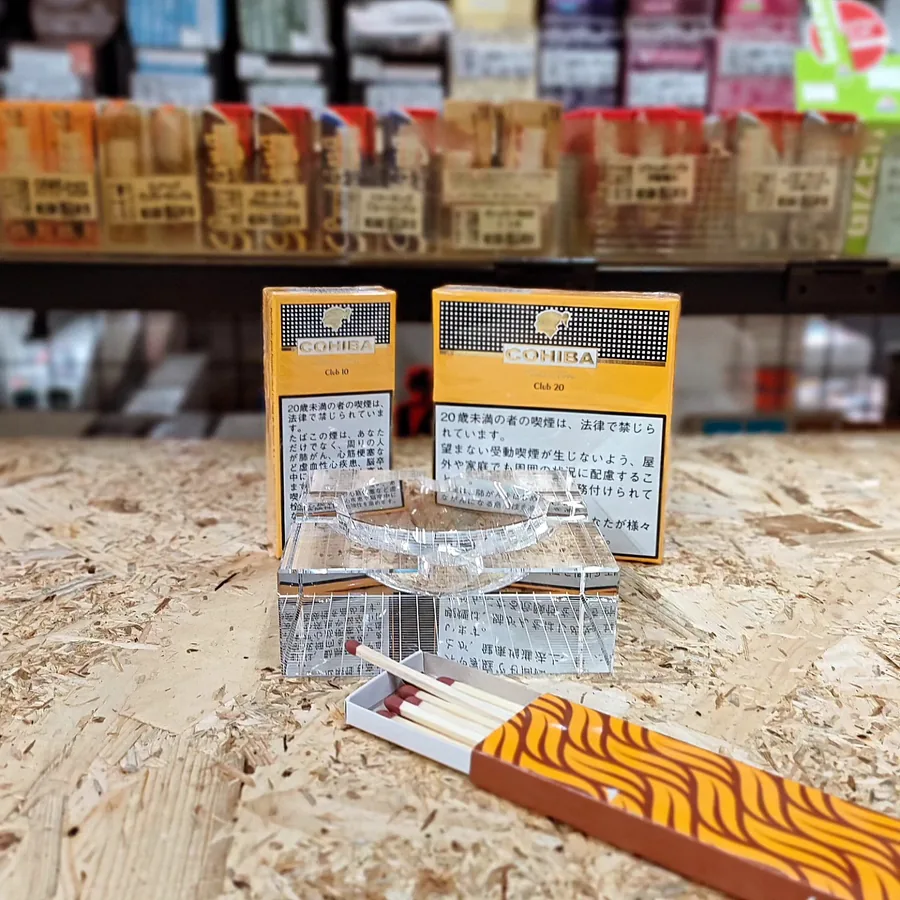Cohiba Club 시가릴로 두 상자(10개입과 20개입)가 유리 재떨이 옆에 놓여 있고, 뒤로는 초점이 나간 가게의 리틀 시가 선반이 보입니다. 2024년 1월 — Art Shobo, Gifu