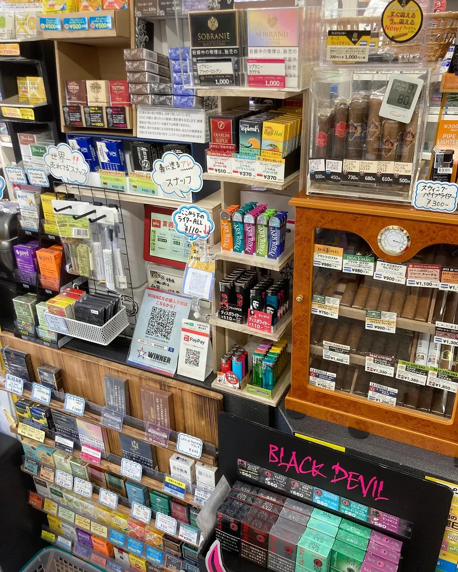 카운터 쪽 진열벽 — Sobranie, Djarum, Black Devil 담배, 독일산 스너프, Ark Royal 라이터, 그리고 버얼 우드 휴미더에 담긴 Don Tomás, Joya de Nicaragua, Macanudo, La Estrella의 낱개 시가 — Blue Lace, Ikoma