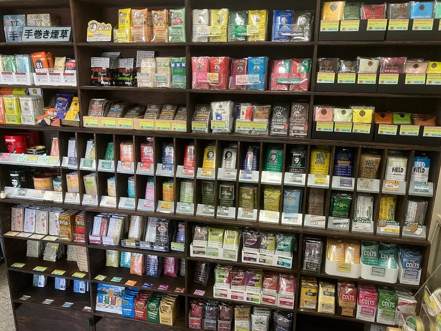 手巻き煙草 간판 아래의 롤링 담배 진열벽 — Ark Royal, Black Spider, Three Dogs, Lumber Jack, Bali Shag, Manitou, Pueblo, Camel, Amber Leaf, Redfield, Choice, Colts — Blue Lace, Ikoma