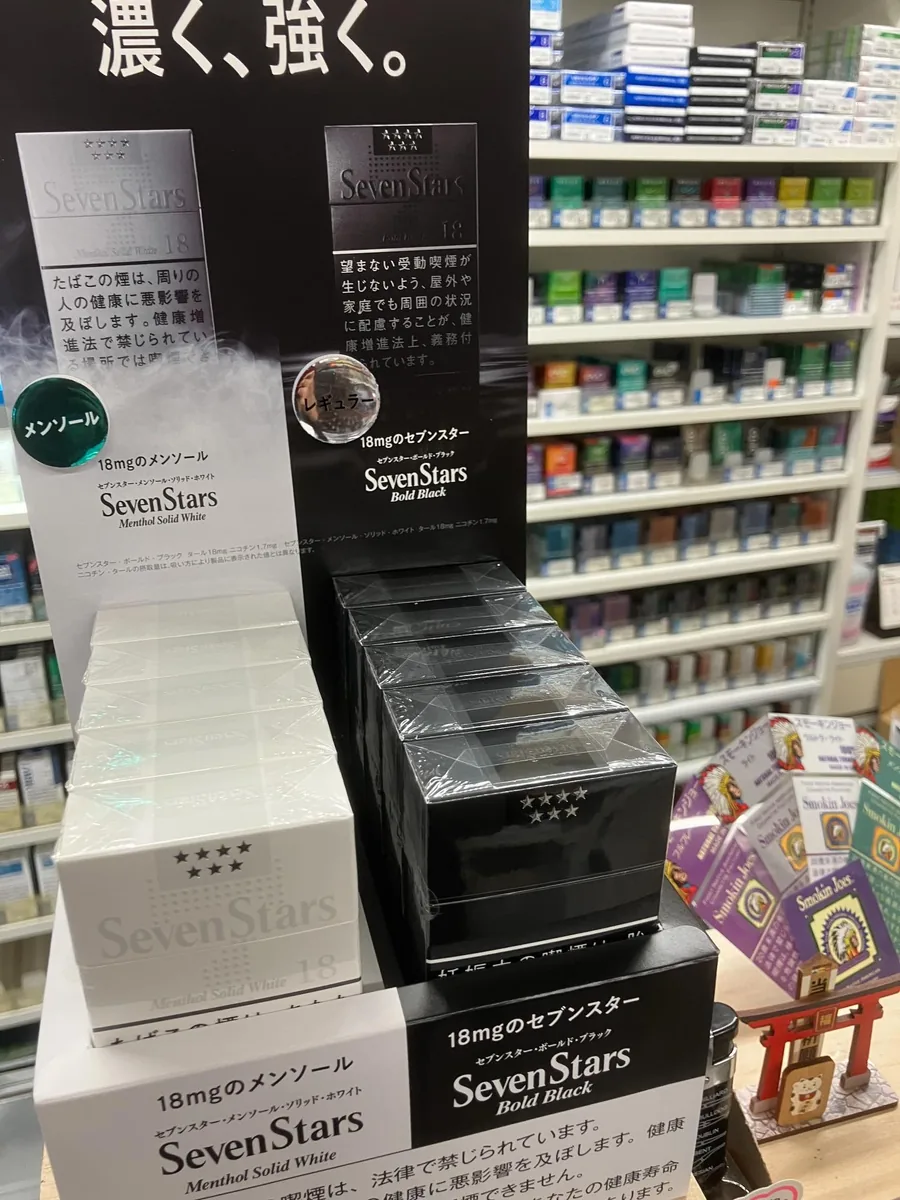 카운터 스탠드에 쌓인 Seven Stars Bold Black과 Seven Stars Menthol Solid White(18 mg), 오른쪽에는 Smokin' Joe's 담배 갑과 작은 도리이·마네키네코 장식 — Blue Lace, Ikoma