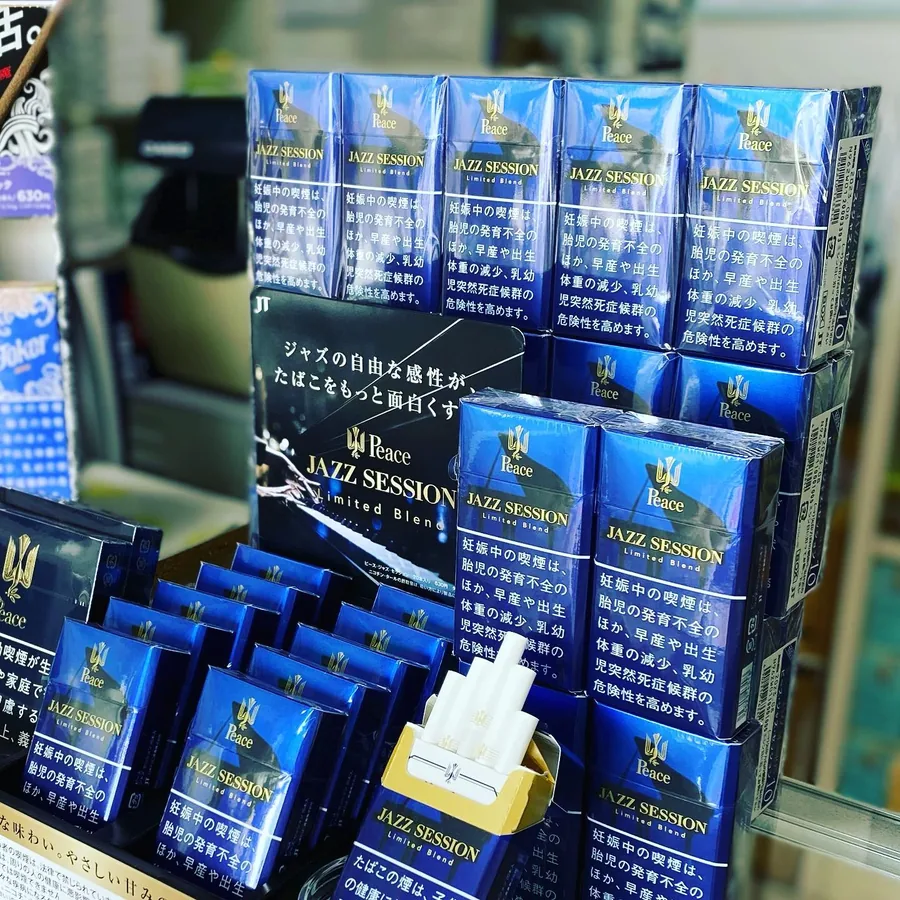 JT 진열 카드 앞에 쌓인 Peace Jazz Session Limited Blend 담배, 한 갑은 개봉된 상태 — Himawari Tobacco Shop, Tsuruga
