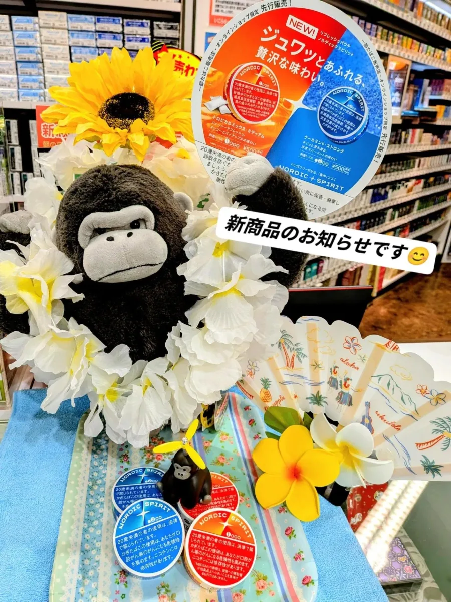 신제품 Nordic Spirit 리프레시 파우치 Tropical Mix (Medium)와 Cool Mint (Strong)를 소개하는 카운터 진열 — 꽃목걸이를 건 고릴라 인형, 해바라기, 종이 부채, Nordic Spirit 틴, 그리고 뒤쪽의 담배 선반 — Icora Mall Tobacco Shop, Izumisano