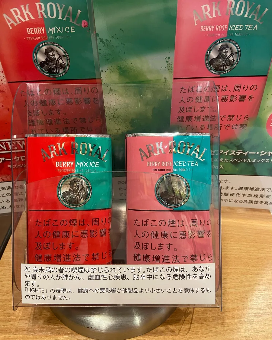 카운터 스탠드에 놓인 Ark Royal Berry Mix Ice와 Berry Rose Iced Tea 롤링 담배, 뒤에는 신제품 포스터 — Tobacco Center Komeda, Gose