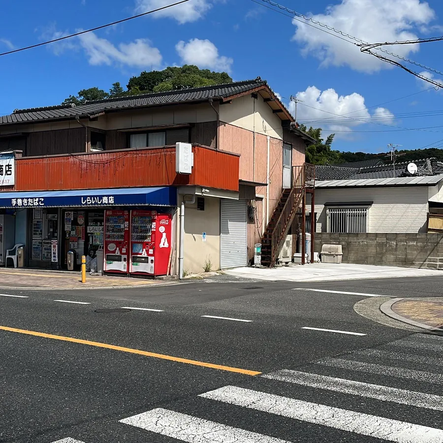 화창한 날 길 건너편에서 본 가게 모퉁이 — 파란 차양, 빨간 코카콜라 음료 자판기 두 대, 2층의 たばこ 간판 — Shiraishi Store, Mizumaki