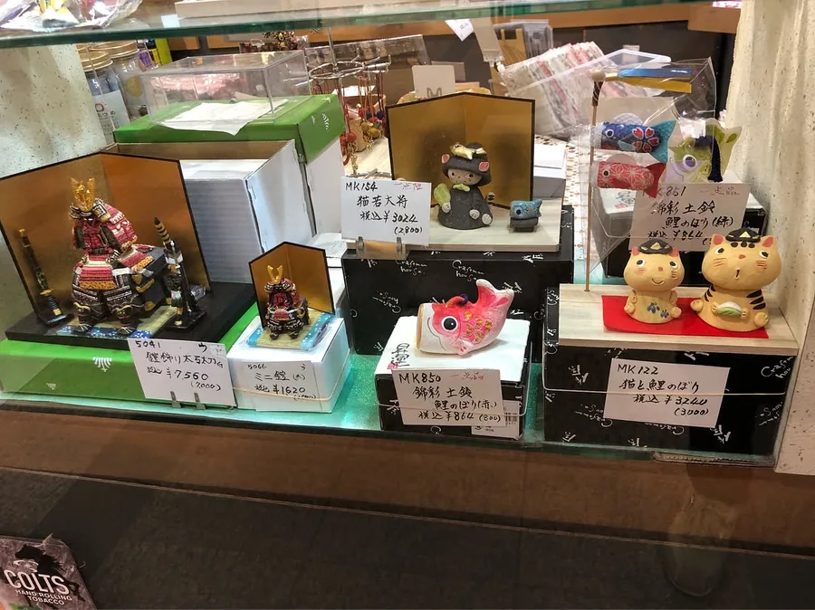 남자아이의 날(端午の節句) 장식을 진열한 쇼윈도 — 무사 갑옷 인형, 잉어 깃발(고이노보리), 호랑이 고양이 장식이 선반 라벨과 함께 놓여 있습니다. 가게의 2018년 4월 게시글은 이를 철 맞춰 내놓는 절기(お節句) 용품이라고 부르며, 하나뿐인 물건이 많다고 소개합니다 — Shop Kita, Kawagoe