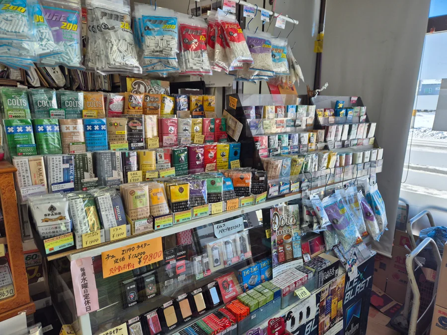 Super Kato Shokai의 롤링 타바코(RYO) 진열대 — Colts, Amsterdamer, Dora, Old Holborn, Ark Royal, Mac Baren, Drum, Spade, Manitou, Golden Virginia 파우치, 위에 걸린 Pure 브랜드 필터, 아래 진열장 속 Che Guevara Zippo와 Zig-Zag 페이퍼, 그리고 유리에 붙인 매장 자필 수요일 휴무 안내문