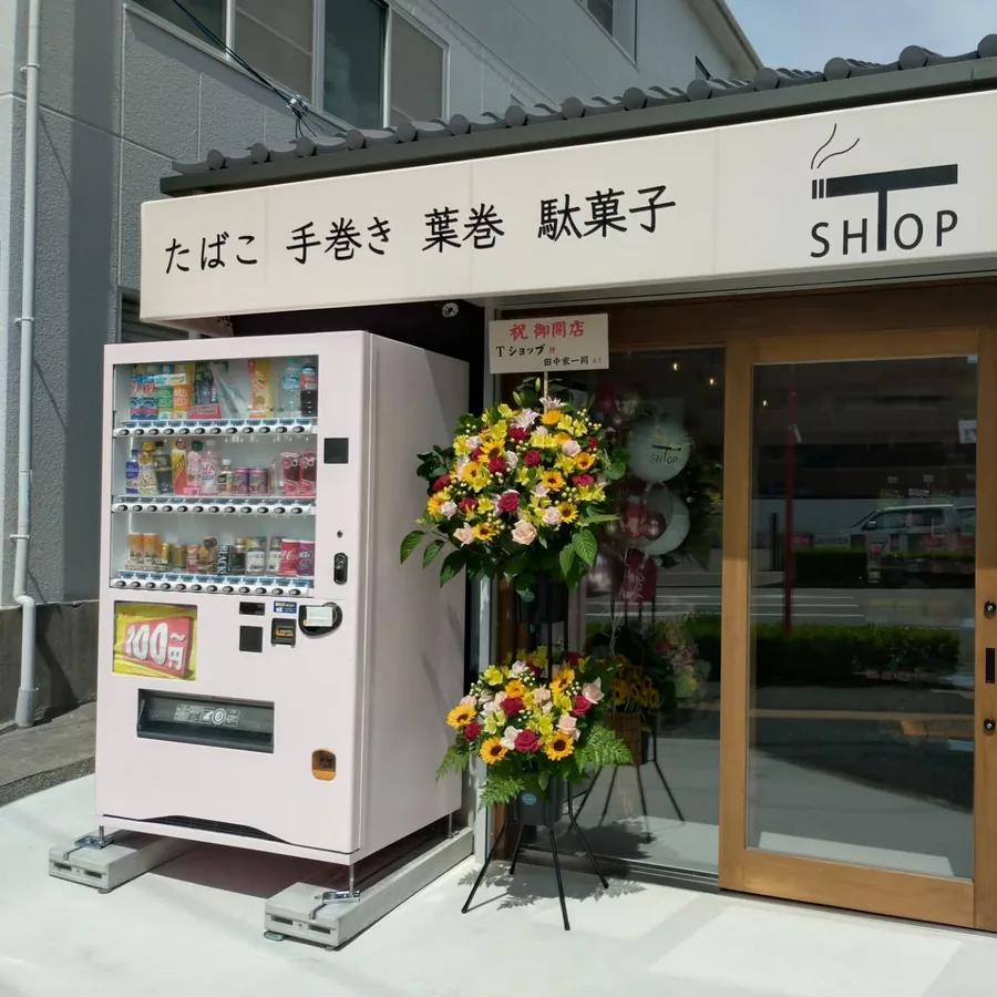 "たばこ 手巻き 葉巻 駄菓子"라고 쓰인 간판 아래, 입구 옆에 놓인 음료 자판기와 문 옆의 개업 축하 화환 — T-Shop Tobacco, Akashi