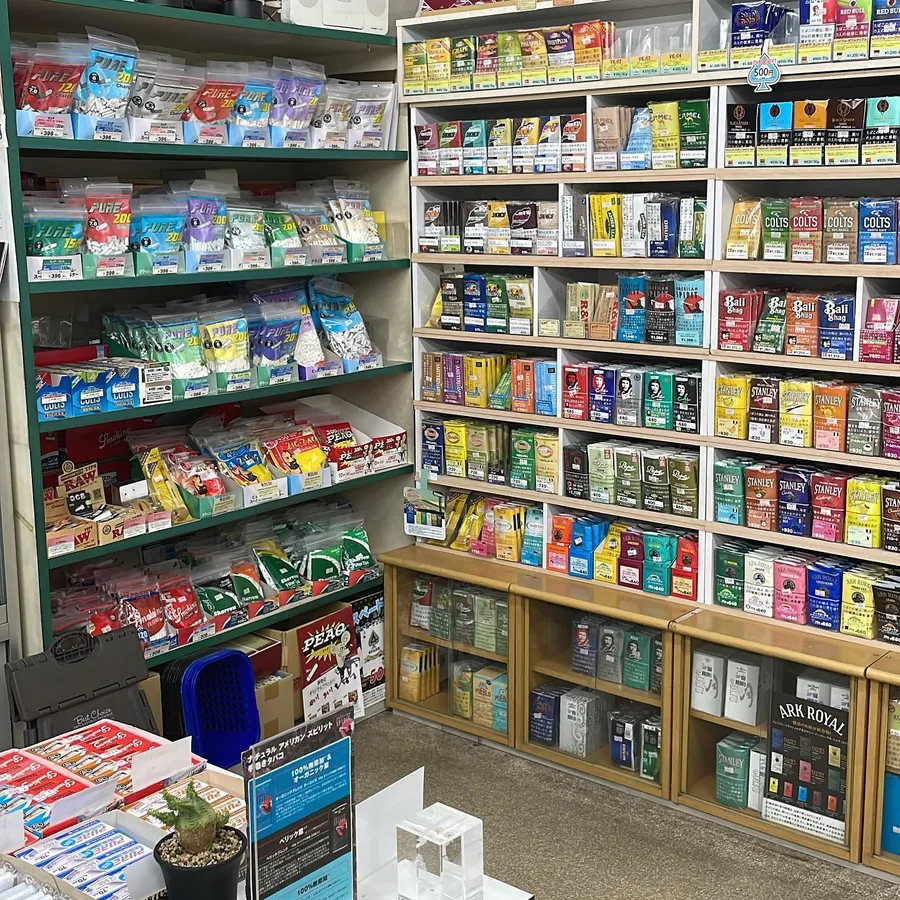 벽 선반에 놓인 롤링 담배 — Choice, Camel, Black Spider, Colts, Bali Shag, American Spirit, Che, Stanley, Pepe, Ark Royal 등 — 그 옆에는 PURE 롤링 필터와 롤링 페이퍼가 놓인 초록색 선반 — Yamada Store, Kasugai